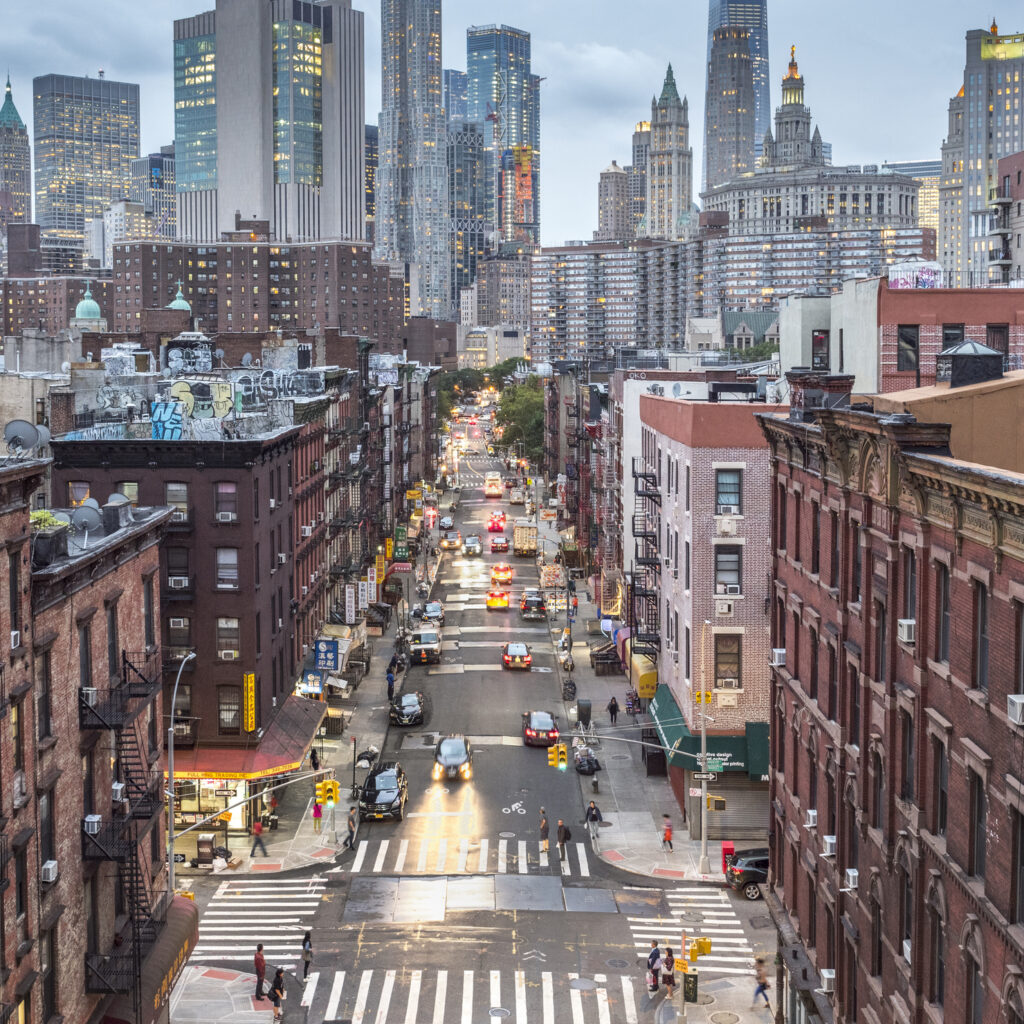 Elevated view of a Lower Manhattan street at dusk, with the New York City skyline rising behind older brick buildings