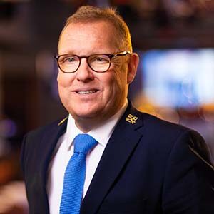 A portrait of Alan Noone. He is a white man with short brown hair, tortoiseshell glasses and a dark suit