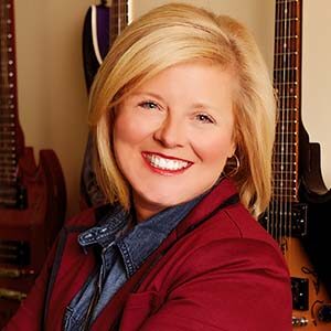 A portrait of Deana Ivey. She is a blonde white woman with shoulder-length hair and a red sweater