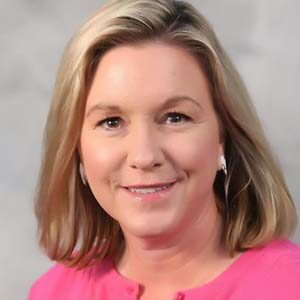 A portrait of Kathy Nelson. She is a white woman with shoulder-length blonde hair and a pink sweater