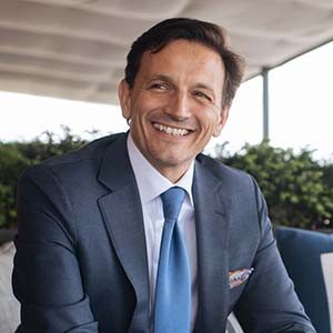 A portrait of Pascal Forotti. He is a tan man with dark combed hair and a blue suit
