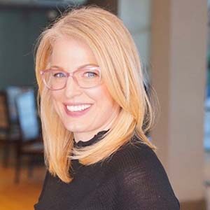 A portrait of Tara Anne Rothberg. She is a blonde white woman with pink glasses and a black sheer blouse