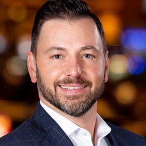 A portrait of Travis Lunn. He is a white man with combed black hair, a stubbled beard and a blue plaid suit