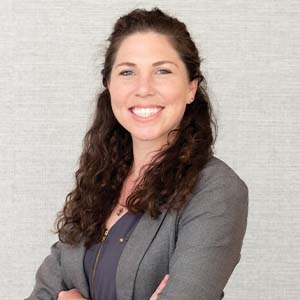 A portrait of Becca Colbert. She is a white woman with long curly brown hair and a grey suit jacket
