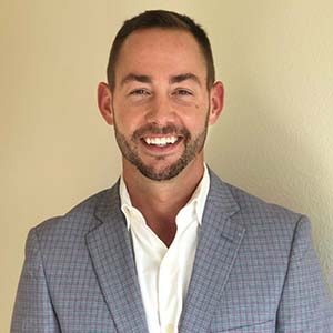 A portrait of Ben Fairchild. He is a white man with short brown hair, a stubbled beard and a plaid grey suit