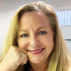 A portrait of Carol M. Smith. She is a white woman with straight blonde hair with one hand at her face