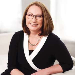 A portrait of Cathy Breden. She is a white woman with shoulder-length brown hair, square glasses and a black blouse with a white collar