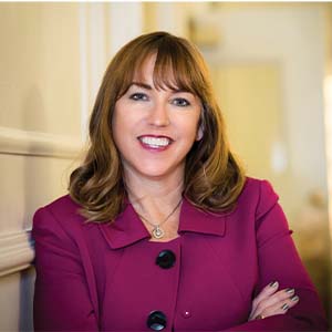 A portrait of Jenn Glynn. She is a white woman with wavy red hair and a pink felt jacket