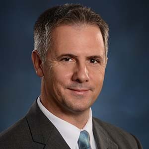 A portrait of Joe Holody. He is a white man with greying combed hair and a brown suit