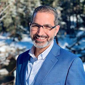 A portrait of John D’Angelo. He is a man with black combed hair, square glasses and a blue suit