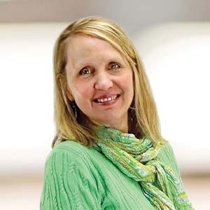 A portrait of Lynn McCullough. She is an older white woman with long blonde hair, a green scarf and a green sweater