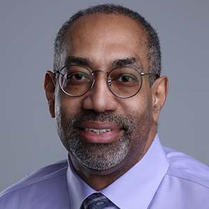 A portrait of Michael Thompson. He is a balding black man with round glasses, a stubbled beard and a blue collared shirt 