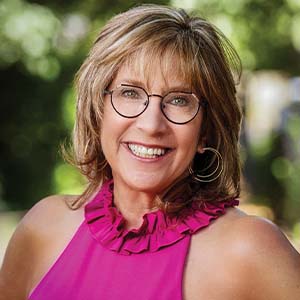 A portrait of Nancy D. Shaffer. She is a white woman with short highlighted hair, round glasses and a pink sleeveless blouse
