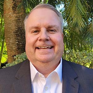 A portrait of Richard Szilagyi. He is a white man with combed grey hair and a grey suit standing in front of palm trees