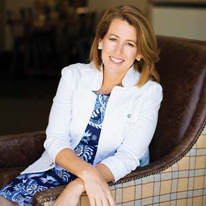 A portrait of Shelly Archer. She is a white woman with highlighted brown hair, a white jacket and a patterned blue dress