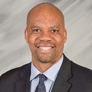 A portrait of William Adams. He is a bald black man with freckles, a stubbled beard and a black suit
