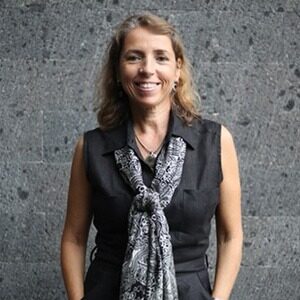 A portrait of Ana Henriques. She is a woman with blonde wavy hair and a sleeveless grey collared shirt