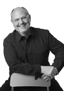 A portrait of Bob Priest-Heck. He is a balding white man with glasses and a black collared shirt