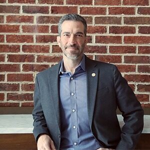 A portrait of Ryan Cornelius. He is an older white man with a black suit jacket and blue collared shirt.