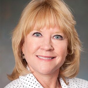 A portrait of Sally Shaffer. She is a white woman with shoulder-length blonde hair and a white blouse.