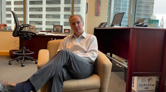 A portrait of Tom Norwalk. He is sitting cross-legged in an armchair with a white collared shirt.