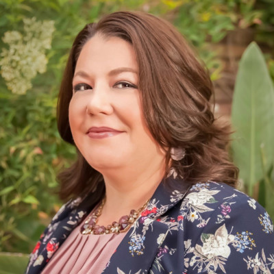A portrait of Abby Hobbs. She is a Native American woman with wavy brown hair and a floral suit jacket.