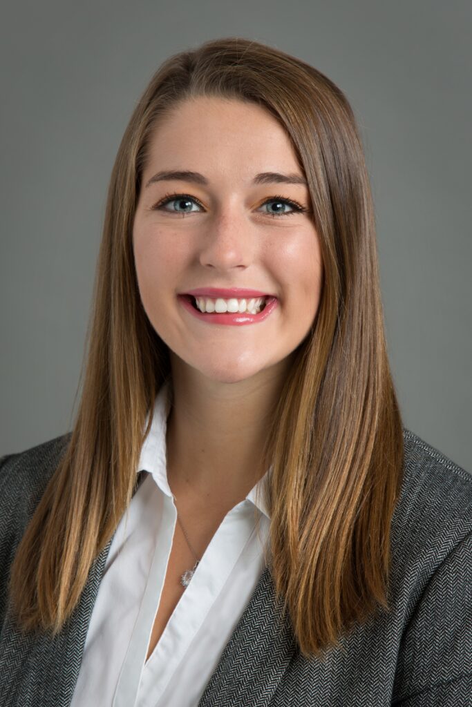 A portrait of Olivia Novak, a young white woman with straight brown hair and a grey suit jacket.