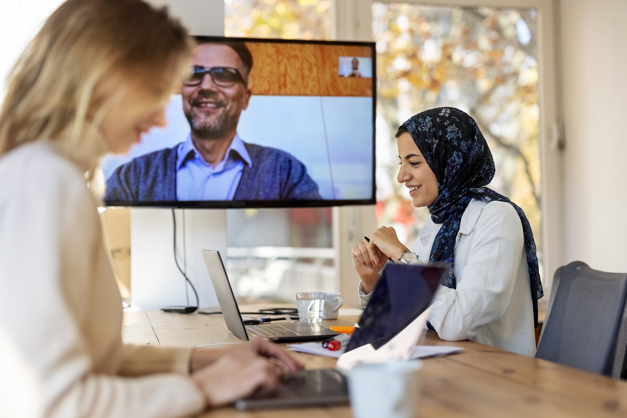 Two women work at a table with laptops while a man speaking through a video call on a screen.