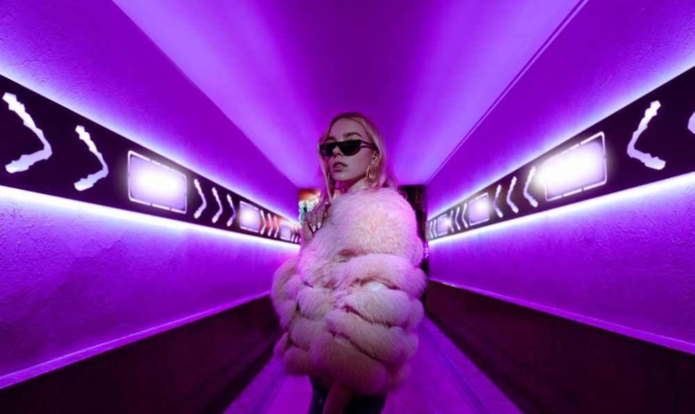 Teen hipster girl in stylish glasses and fur standing in purple neon lights on street wall