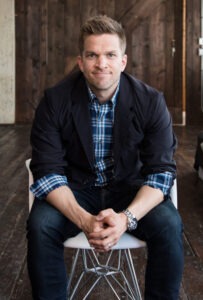 man sitting down, wearing black coat and blue plaid long sleeve button up shirt