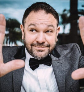 man holding up hands to camera, wearing black bowtie, dark grey sport coat and white dress shirt