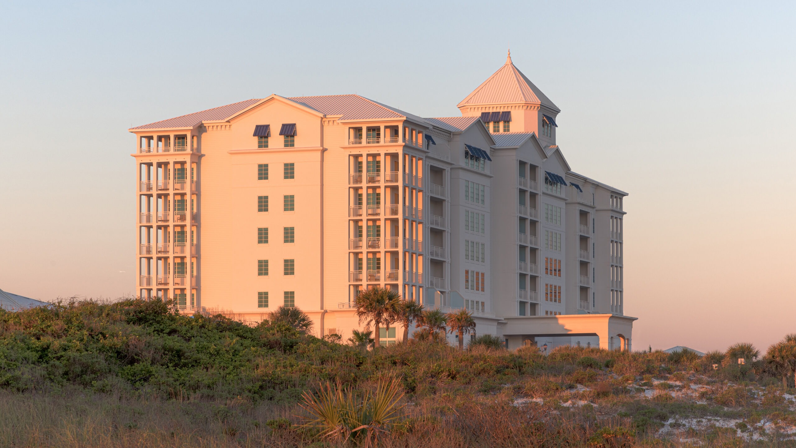 A multi-story resort stands on the beach, reflecting orange light from the setting sun