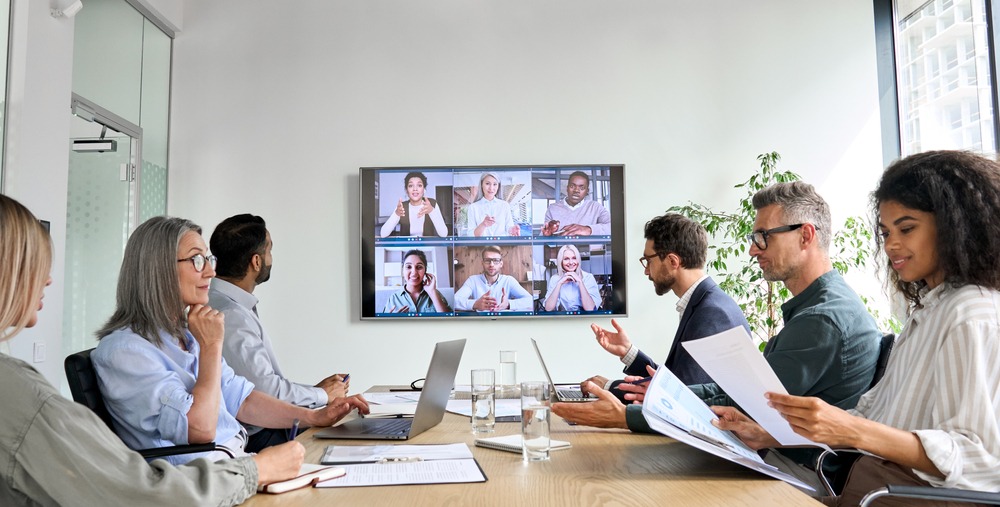 picture of board room with diverse group of people looking at paper and laptops and Zoom faces on big screen