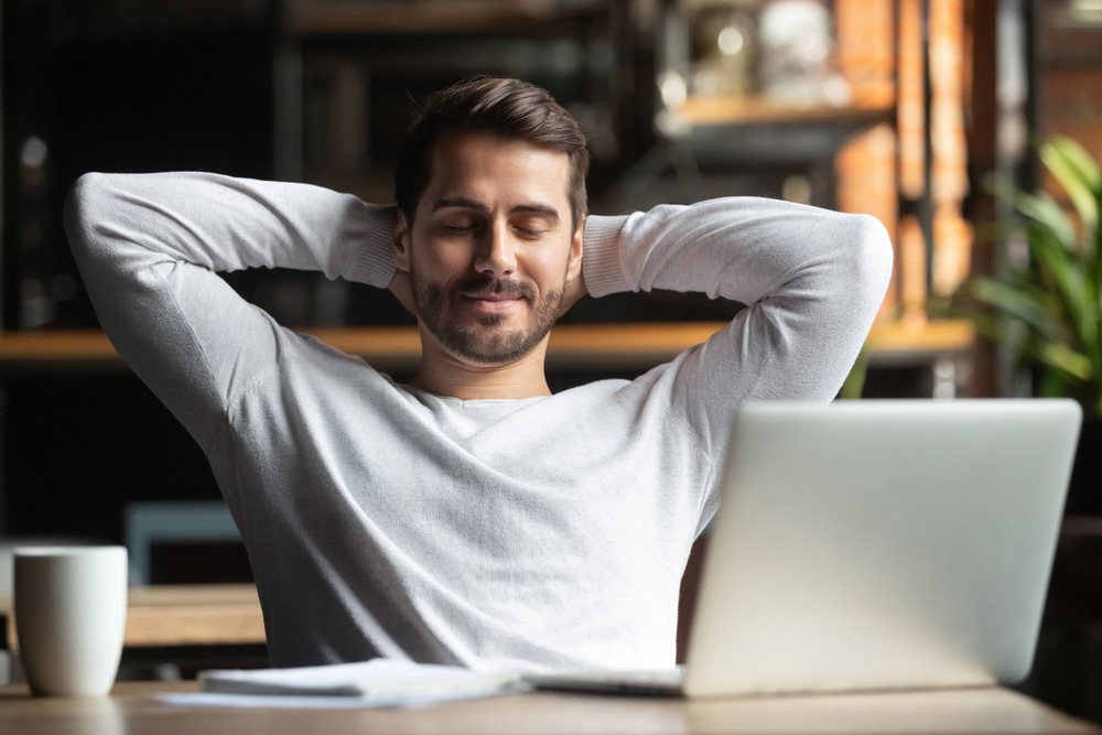 man with eyes closed sitting in front of computer with hands behind head